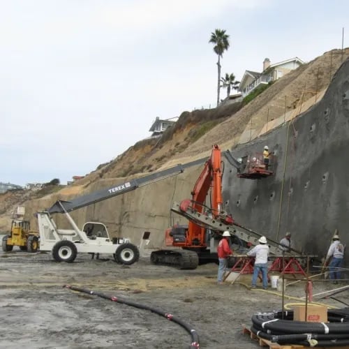 Heavy machinery works on the sand of a beach at low tide carving away and covering the bluff with concrete.